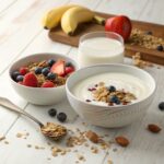 A breakfast setup featuring two cereal bowls—one with milk and one with yogurt—topped with fresh berries, banana slices, and nuts, placed on a rustic wooden table.