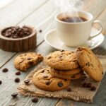 Freshly baked coffee cookies with chocolate chips and coffee beans on a rustic wooden table alongside a steaming cup of coffee.