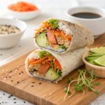 A sushi burrito cut in half showing layers of brown rice, vegetables, avocado, and grilled salmon, served with soy sauce and garnishes.